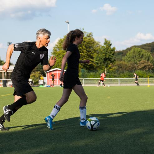 Trainer joggt hinter einem M&auml;dchen, das einen Fu&szlig;ball auf einem gr&uuml;nen Sportplatz dribbelt, im Hintergrund sind weitere Spieler.