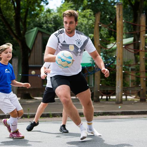 Junger Mann in Sportswear spielt Fu&szlig;ball mit einem M&auml;dchen und einem weiteren Jungen auf einem Platz im Freien.