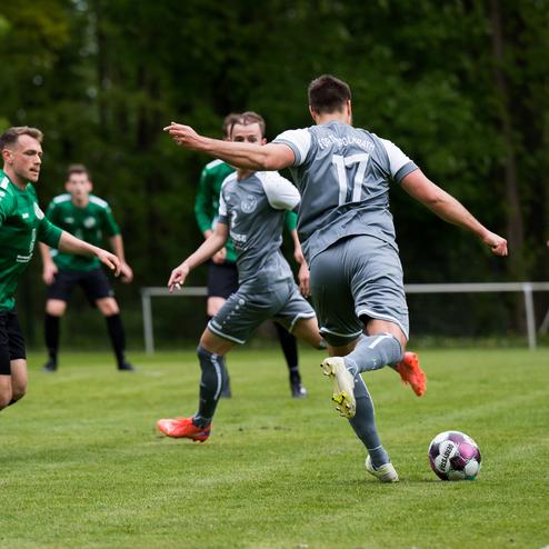 Spielszene mit Fu&szlig;ballspielern, einer in grauem Trikot mit Ball, zwei Gegnern in gr&uuml;nen Trikots im Hintergrund.