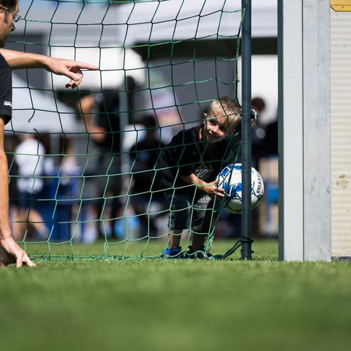 Ein Kind schaut durch das Fu&szlig;balltor und h&auml;lt einen Ball, w&auml;hrend ein Erwachsener auf es zeigt.
