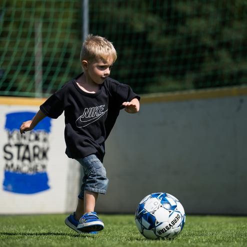 Ein kleiner Junge l&auml;uft spielerisch mit einem Fu&szlig;ball auf einem gr&uuml;nen Platz in einem Tor alle.