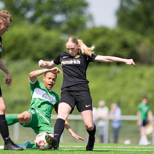 Zwei Fu&szlig;ballspielerinnen k&auml;mpfen um den Ball auf einem Rasenplatz; ein Spieler liegt am Boden.