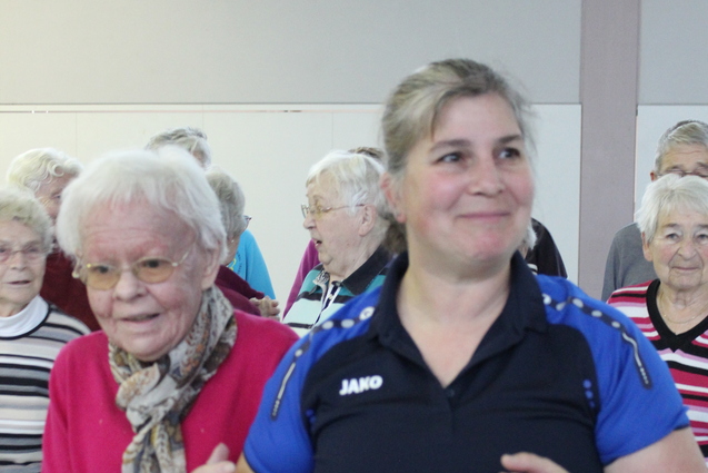 Eine Seniorenbetreuerin f&uuml;hrt eine &auml;ltere Dame beim Tanzen in einer Gruppe von &auml;lteren Menschen.