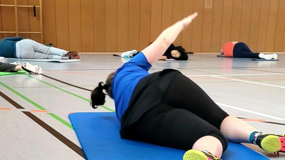 Person in blauer Sportkleidung liegt auf einer Yogamatte und führt eine Übung in einer Sporthalle durch.