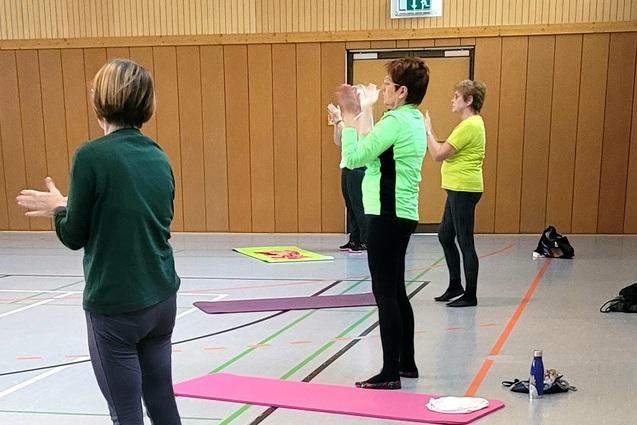 Gruppentraining in einer Halle, mit Teilnehmern auf Matten, die aktiv &Uuml;bungen durchf&uuml;hren und applaudieren.