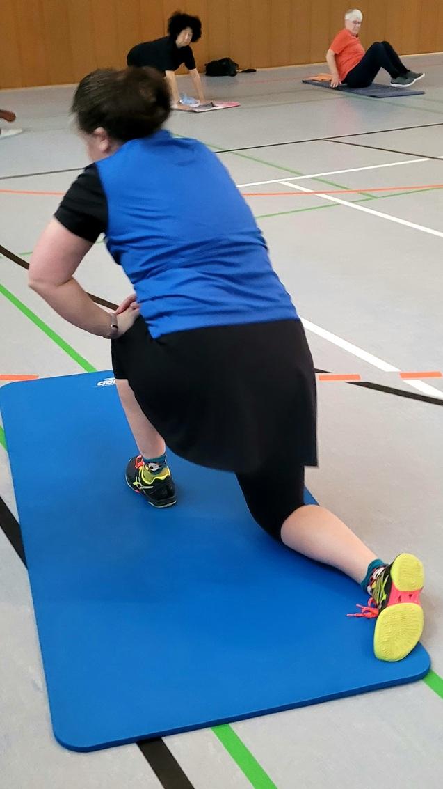 Frau in blauer Sportbekleidung dehnt sich auf einer blauen Matte in einer Sporthalle, im Hintergrund weitere Personen.