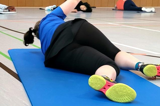 Person in blauer Sportkleidung liegt auf einer Yogamatte und f&uuml;hrt eine &Uuml;bung in einer Sporthalle durch.