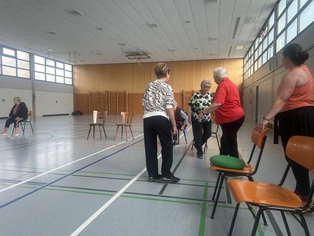 Begrüßung Senioren bei einem Übungsangebot in einer Sporthalle, einige stehen, andere sitzen auf Stühlen.