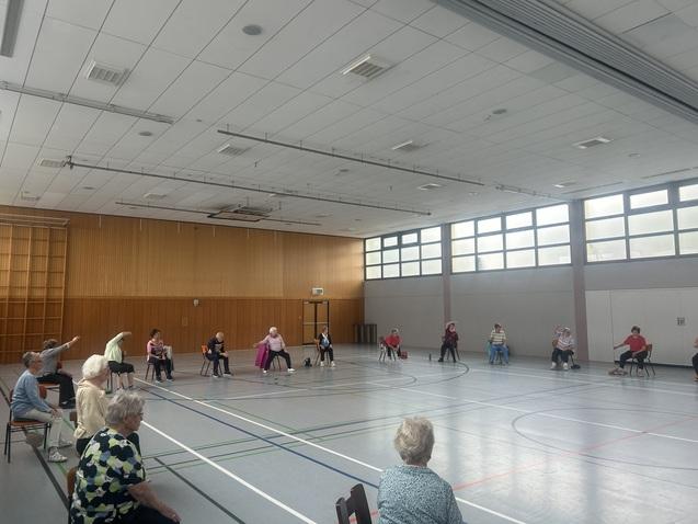 Ältere Personen sitzen in einem großen Raum auf Stühlen und machen Bewegungsübungen zusammen.