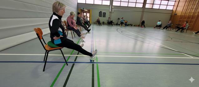 Senioren sitzen auf Stühlen in einer Sporthalle, einige führen Beinübungen durch, andere beobachten.