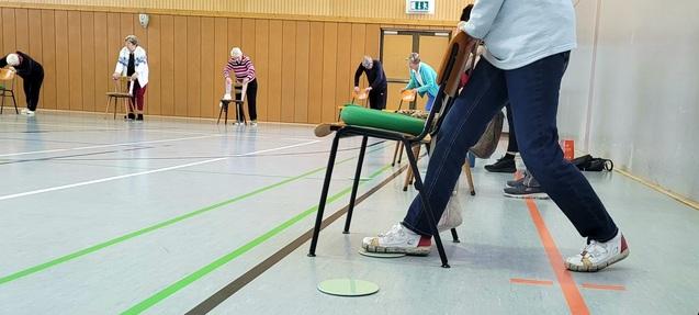 Gruppenübung mit Stühlen in einer Sporthalle; ältere Menschen aktivieren sich und dehnen sich.