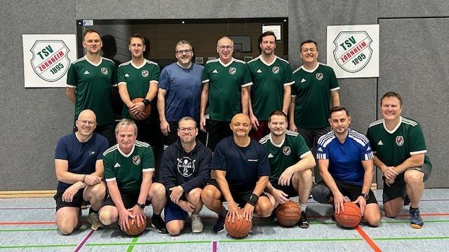 Gruppe von elf Basketballspielern in Sportkleidung mit B&auml;llen in einer Turnhalle, vor der Wand mit Vereinslogos.