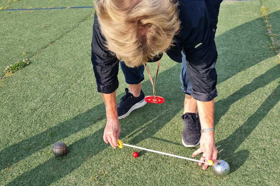 Person misst den Abstand zwischen einem roten und einem silbernen Kugel auf einem Rasenplatz mit einem Maßband.