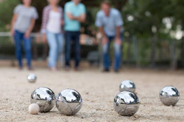 Nahaufnahme von metallischen Boule-Kugeln und einer Holz-Kugel auf sandigem Boden, während Personen im Hintergrund zuschauen.