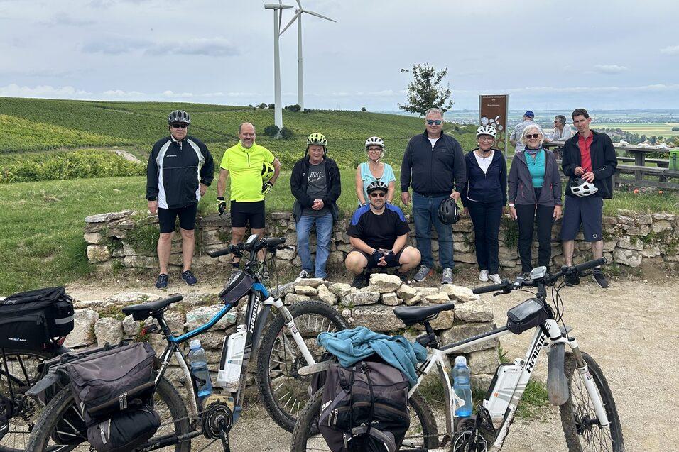 Gruppe von Radfahrern posiert vor Windr&auml;dern und gr&uuml;nem Landschaftshorizont, Fahrr&auml;der liegen vor ihnen am Boden.