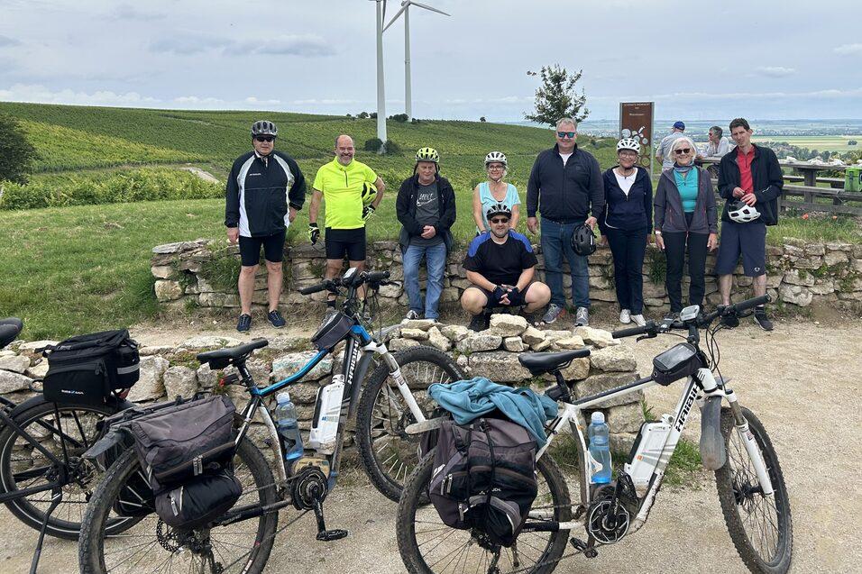 Gruppe von Radfahrern posiert vor Windr&auml;dern und gr&uuml;nem Landschaftshorizont, Fahrr&auml;der liegen vor ihnen am Boden.
