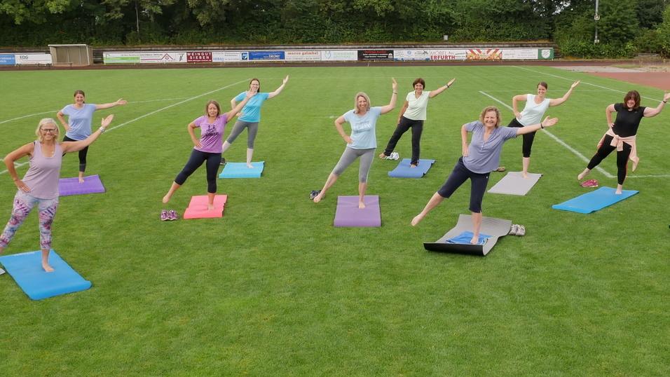 Gruppe von zehn Frauen in Fitnesskleidung, die auf Matten auf einem Sportplatz &Uuml;bungen machen.