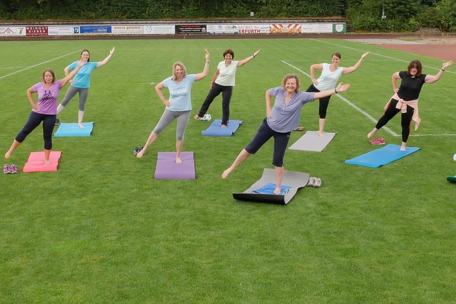Gruppe von zehn Frauen in Fitnesskleidung, die auf Matten auf einem Sportplatz &Uuml;bungen machen.