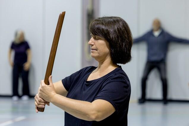 Frau mit Holzstab in der Hand, in einer Sporthalle, w&auml;hrend im Hintergrund andere Personen &Uuml;bungen machen.