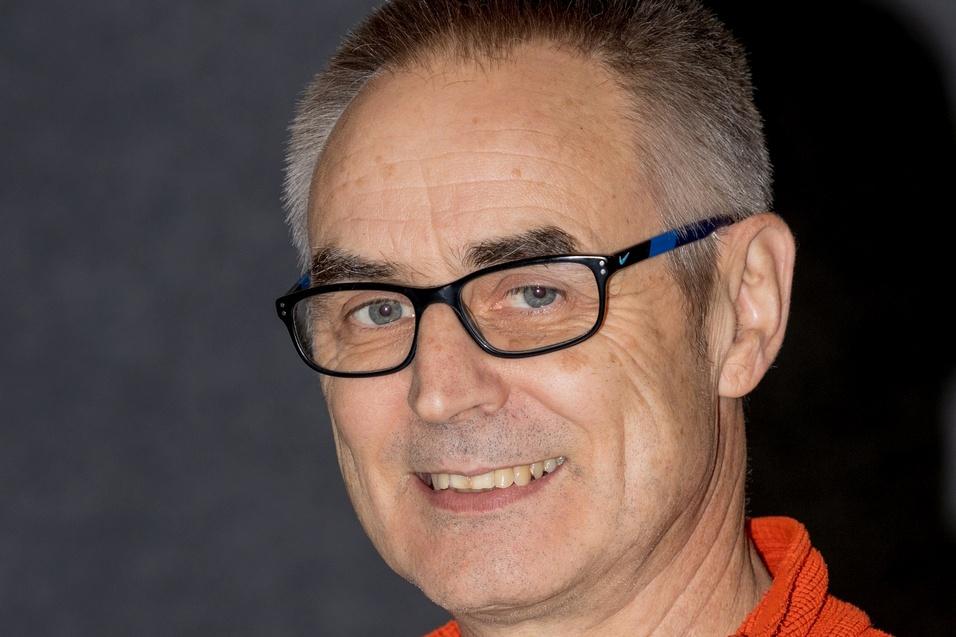 Mann mit Brille tr&auml;gt einen orangefarbenen Fleece-Pullover und l&auml;chelt vor einem dunklen Hintergrund.