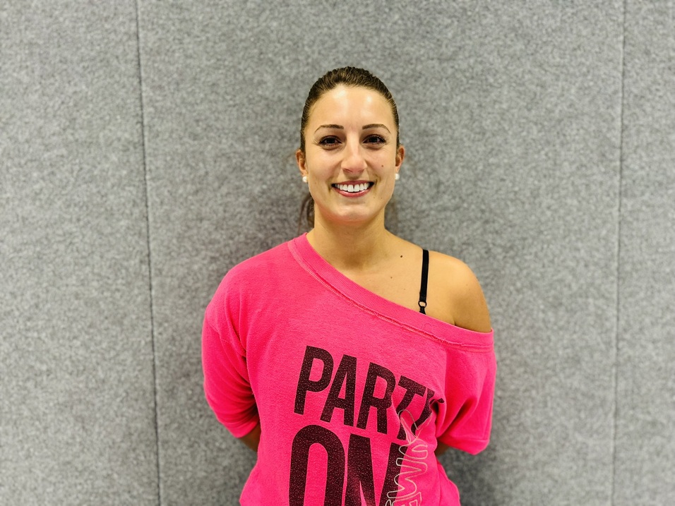 Junge Frau mit hochgestecktem Haar tr&auml;gt ein pinkes, l&auml;ssiges Shirt und l&auml;chelt vor einer grauen Wand.