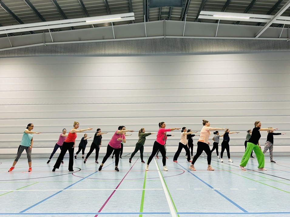 Eine Gruppe von Frauen tanzt synchron in einer Sporthalle mit farbigen Linien auf dem Boden.
