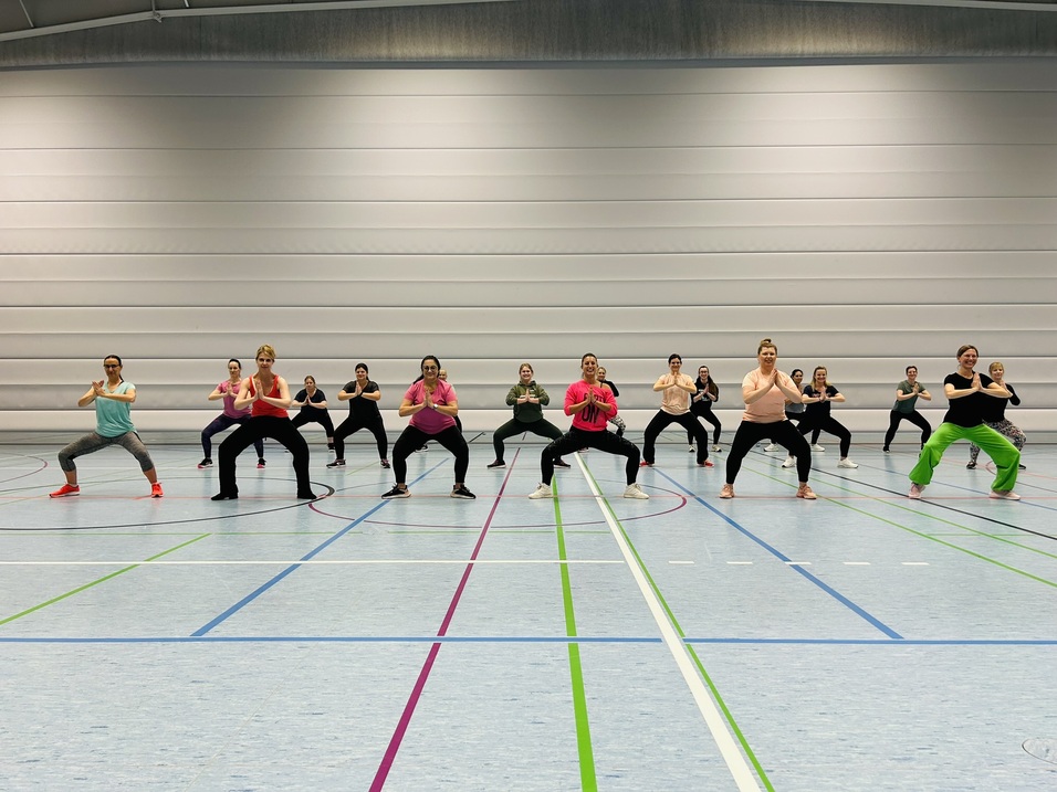 Eine Gruppe von Frauen in Sportkleidung trainiert synchron in einer Sporthalle auf einem bunten Hallenboden.