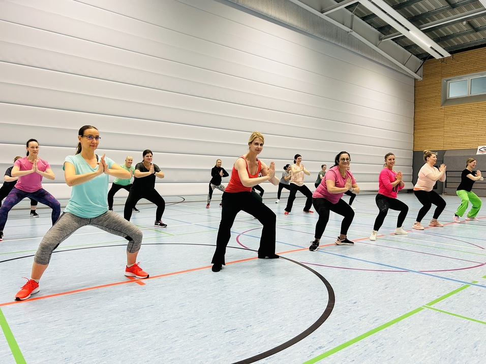 Gruppe von Frauen in Sportkleidung beim Gruppentraining in einer Sporthalle, unterschiedliche Bewegungspositionen.