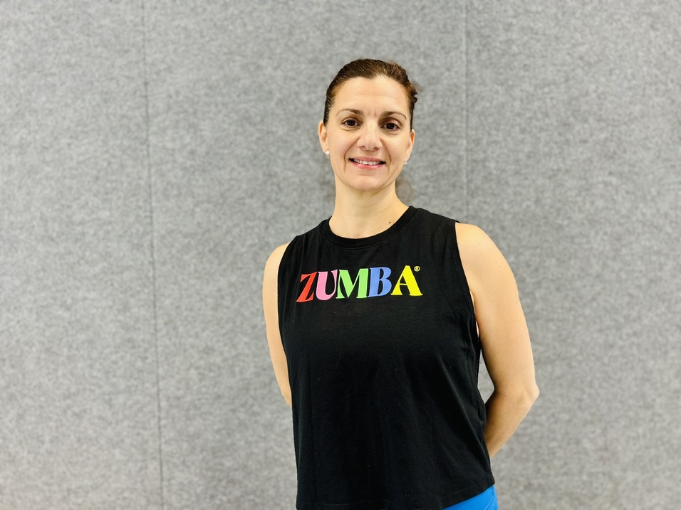 Frau in schwarzer Zumba-Tanktop steht l&auml;chelnd vor grauer Wand und pr&auml;sentiert eine sportliche Haltung.