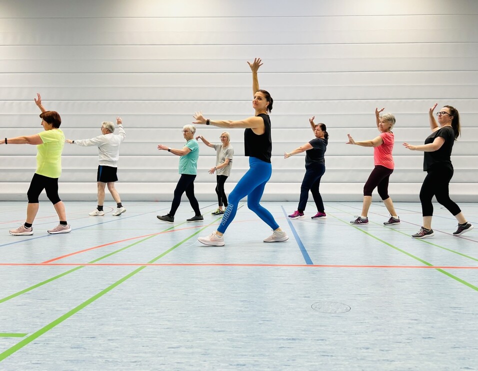 Gruppe von Frauen in Sportbekleidung, die in einer Turnhalle gemeinsam tanzen und Bewegungen synchron ausf&uuml;hren.