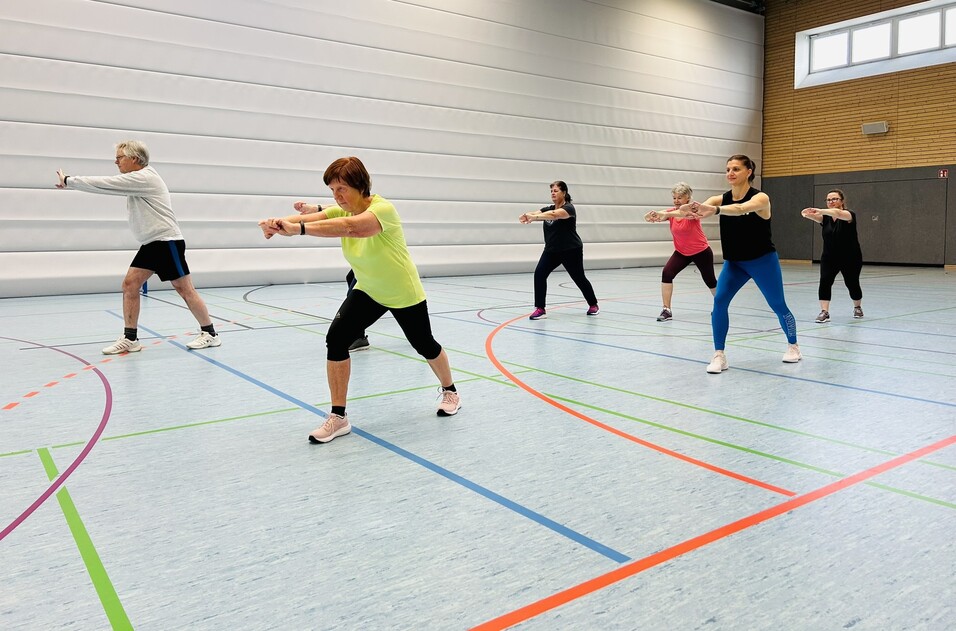 Gruppe von sechs Personen beim Workout in einer Sporthalle, die synchron &Uuml;bungen ausf&uuml;hren.