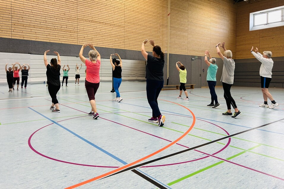 Eine Gruppe von Frauen &uuml;bt synchronen Tanz in einer Sporthalle mit mehrfarbigen Linien auf dem Boden.