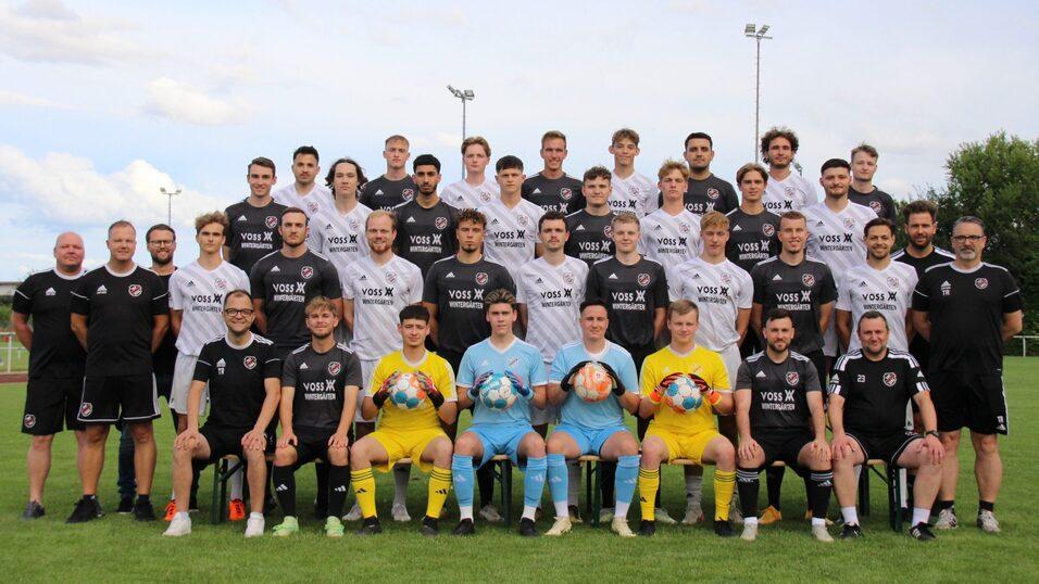 Mannschaftsfoto einer Fu&szlig;ballmannschaft mit Trainern, Spielern in Trikots und Torwarten auf einem Sportplatz.