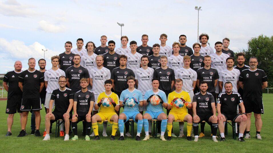 Mannschaftsfoto einer Fu&szlig;ballmannschaft mit Trainern, Spielern in Trikots und Torwarten auf einem Sportplatz.