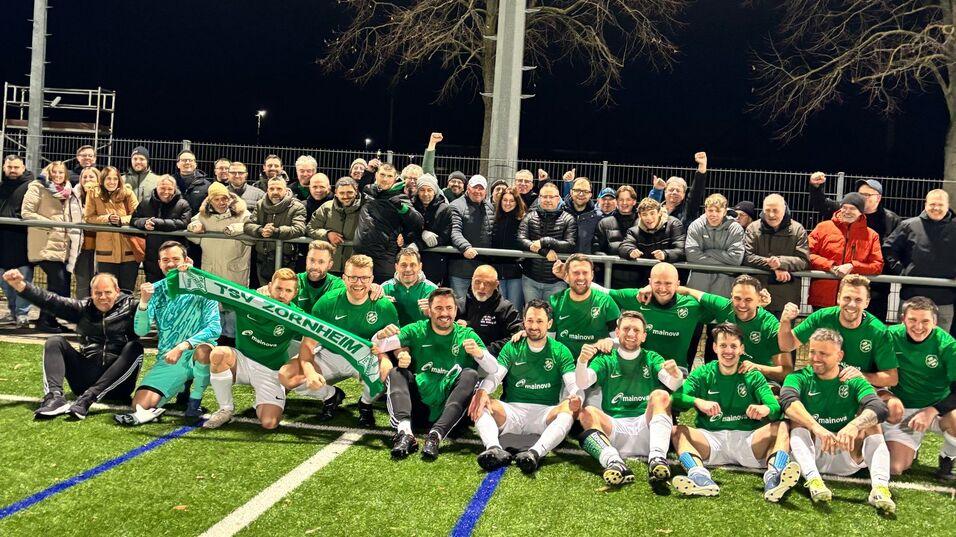 Mannschaftsfoto von Fu&szlig;ballspielern in gr&uuml;nen Trikots mit Fans im Hintergrund auf einem Kunstrasenplatz.
