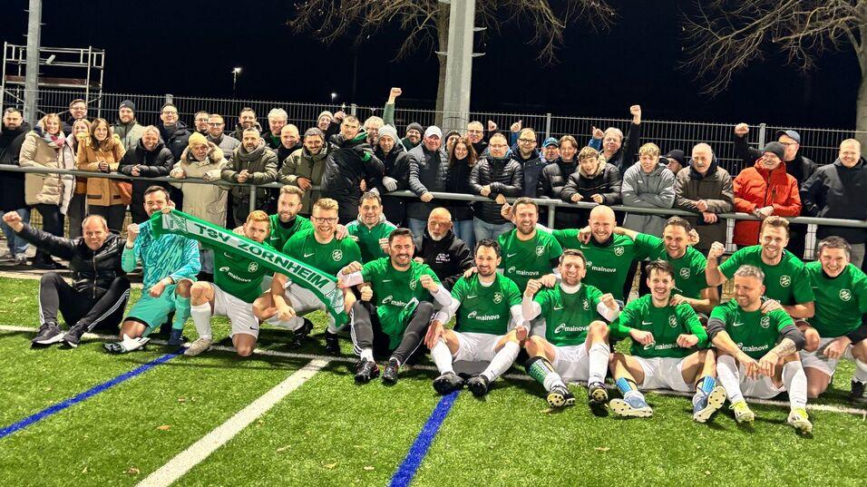 Mannschaftsfoto von Fu&szlig;ballspielern in gr&uuml;nen Trikots mit Fans im Hintergrund auf einem Kunstrasenplatz.