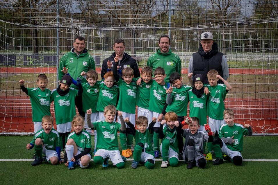Gruppennfoto von Kindern in gr&uuml;nen Fu&szlig;balltrikots mit Trainern auf einem Sportplatz vor einem Tor.