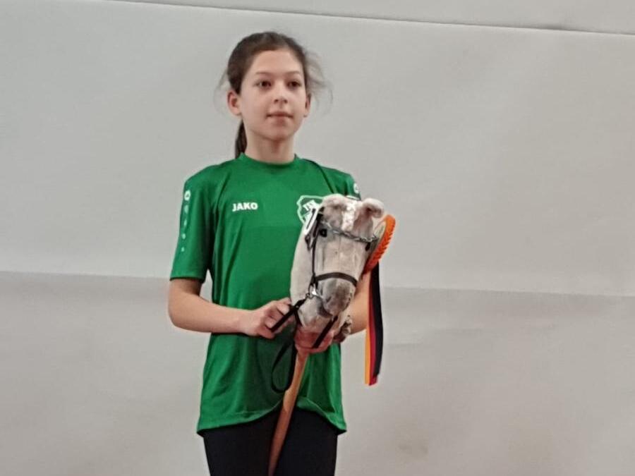 M&auml;dchen in gr&uuml;nem T-Shirt steht auf einem Podest und h&auml;lt ein Stoffpferd mit einer Schleife in den H&auml;nden.