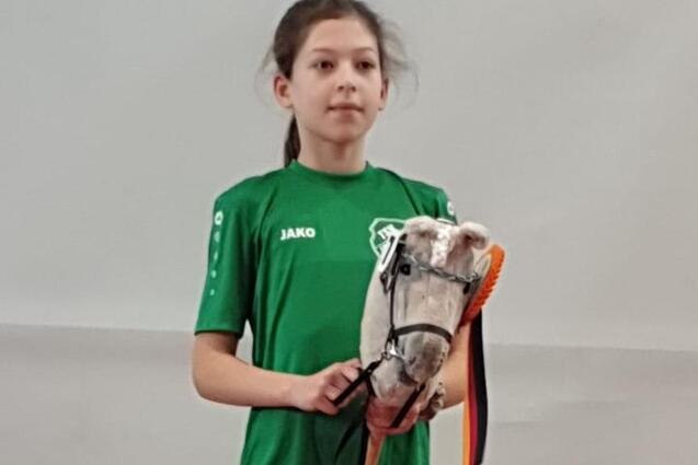 M&auml;dchen in gr&uuml;nem T-Shirt steht auf einem Podest und h&auml;lt ein Stoffpferd mit einer Schleife in den H&auml;nden.