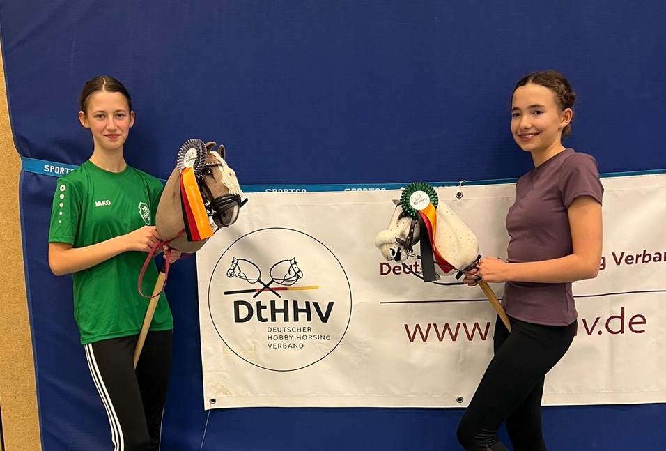 Zwei junge M&auml;dchen halten Pferde-Spielzeugfiguren vor einem Banner des Deutschen Pferdesportverbands in einer Sporthalle.
