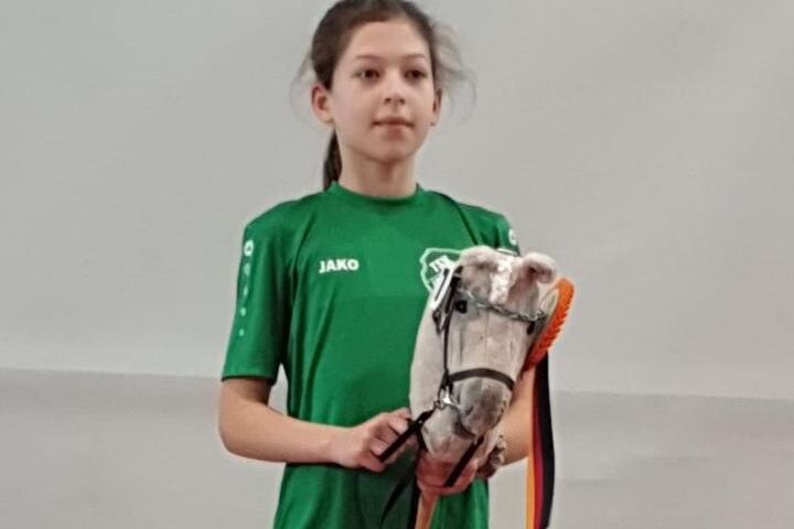 M&auml;dchen in gr&uuml;nem T-Shirt steht auf einem Podest und h&auml;lt ein Stoffpferd mit einer Schleife in den H&auml;nden.