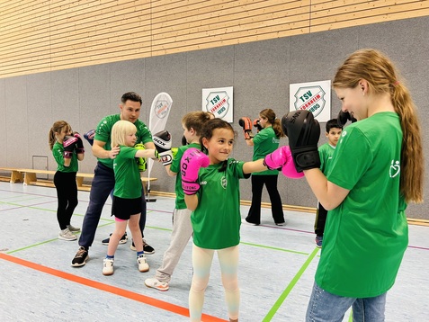 Kinder in gr&uuml;nen T-Shirts trainieren Boxen, w&auml;hrend sie sich gegenseitig mit Handschuhen anfeuern.