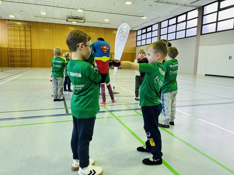 Kinder in gr&uuml;nen T-Shirts &uuml;ben Kampfsporttechniken in einer Sporthalle, w&auml;hrend eine Superheldenfigur im Hintergrund steht.