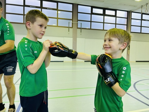 Zwei Jungen in gr&uuml;nen T-Shirts tragen Boxhandschuhe und &uuml;ben Boxbewegungen in einer Sporthalle.