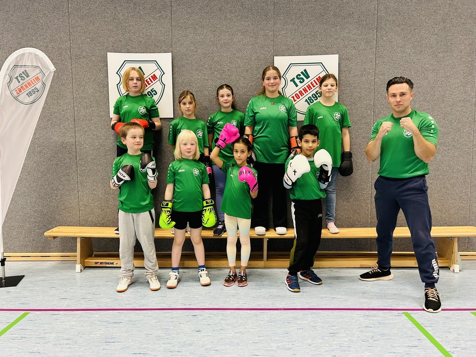 Eine Gruppe von zehn Kindern und einem Trainer in gr&uuml;nen T-Shirts, in Boxhandschuhen, posiert in einer Sporthalle.