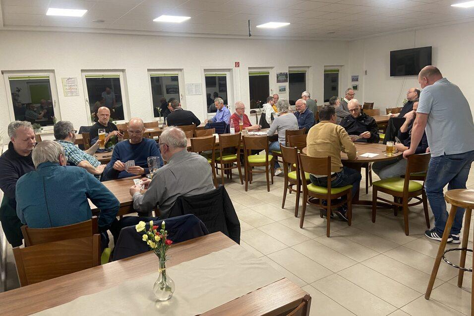 Gruppenszene in einem Versammlungsraum mit M&auml;nnern, die an Tischen sitzen und sich unterhalten.