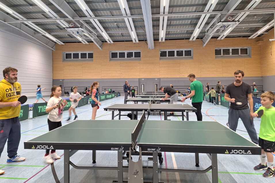Gruppe von Spielern unterschiedlichen Alters beim Tischtennis in einer Sporthalle mit mehreren Tischtennisplatten.