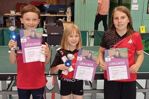 Drei Kinder im Sportoutfit halten Urkunden und Medaillen in einer Tischtennishalle. Tischtennistisch im Hintergrund.