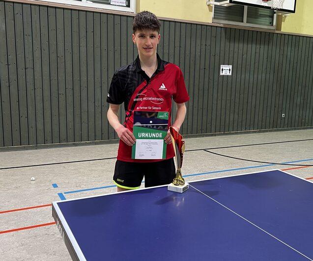 Jugendlicher in rot-schwarz gestreifter Sportkleidung h&auml;lt Urkunde und Pokal vor einem Tischtennistisch.