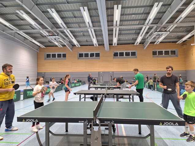 Gruppe von Spielern unterschiedlichen Alters beim Tischtennis in einer Sporthalle mit mehreren Tischtennisplatten.
